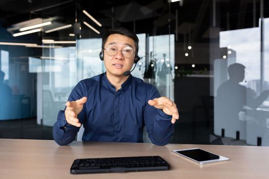 A young Asian man sits in the office at a desk in a headset, looks and talks to the camera. Explains, advises, conducts training, online meeting.
