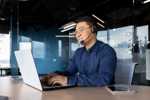 Young male Asian teacher is lecturing students, taking exams online via laptop video call. Sitting in the office at the table in a headset, smiling at the camera.