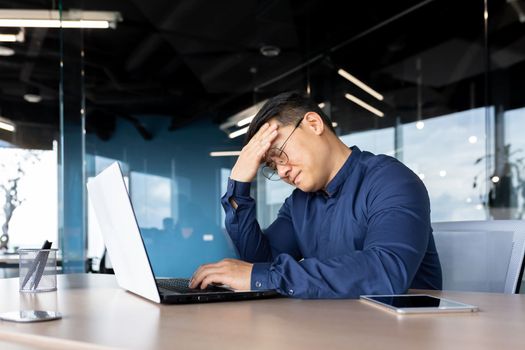 A tired young Asian man sits in the office at the table, holds his head, feels a headache, tension, migraine after a hard overtime day, shakes.