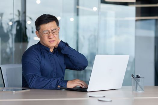 A tired young man, a freelancer, a businessman, sits behind a desk in the office, feels unbearable pain in his neck, closes his eyes, grimaces in pain, massages his hand.