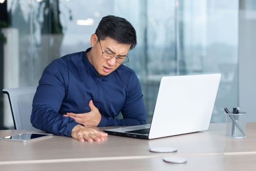 An Asian man has a heart attack, stroke, panic attack, pneumonia at the workplace. He is sitting at the desk in the office, holding his chest with his hands, feeling severe pain, and needs medical help.