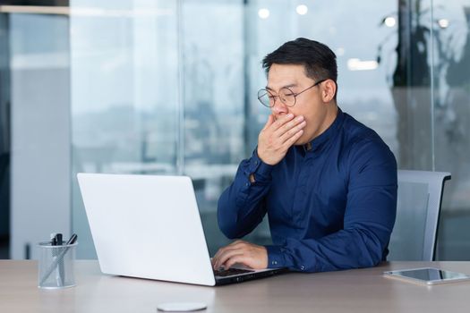 Tired young asian businessman yawns at workplace. He is sitting sleepily at the desk in the office, working overtime at the laptop.