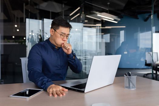 A young Asian businessman, lawyer fell ill at work. sits at the work table. coughs, covers his mouth with his hand, feels bad.