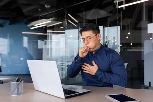 A young Asian man coughs in the office at the desk. Has allergies, flu, asthma. He holds his chest, covers his mouth.