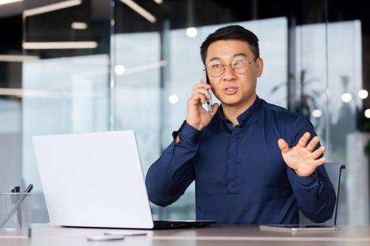 Young man, Asian businessman, student, talking on the phone with clients and partners. Sitting in the office at the table, working on the laptop.