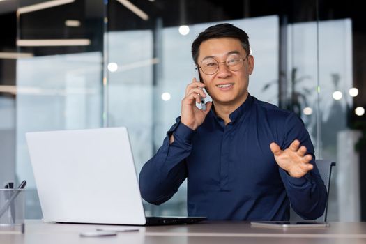 A young Asian man is talking on the phone in the office. He sits at a desk with a laptop, looks at the camera, and smiles.