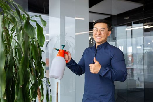 Young male office worker, programmer, freelancer. Asian takes care of plants. Waters and sprays them. He looks at the camera, smiles, points with a super finger.