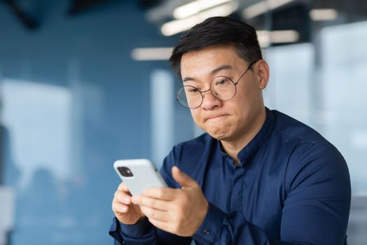 Close-up photo. An angry young Asian man in glasses is holding a phone in his hands. Nervously pursed lips, reads news, messages, loses in online games.