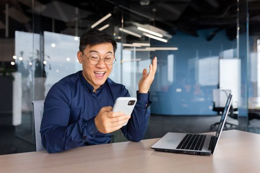 Happy young man businessman, freelancer sitting in the office at the table, holding the phone in his hands. Gladly reads the message, good news.