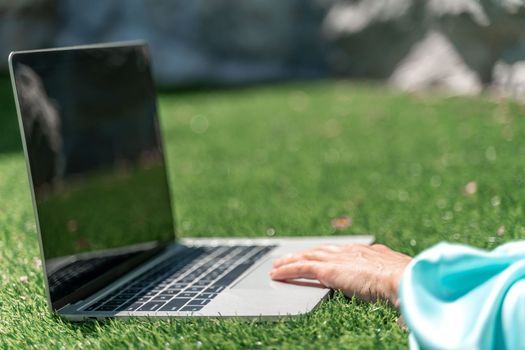 a young beautiful woman with blond curly hair in glasses and a blue dress sits on the grass in nature and uses a laptop