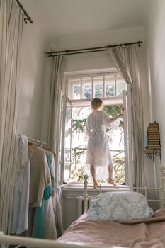 Lifestyle portrait of a lady having fun in a sunlit room. Charming girl with blond hair, cream dress, standing on the windowsill, smiling and looking into the distance.