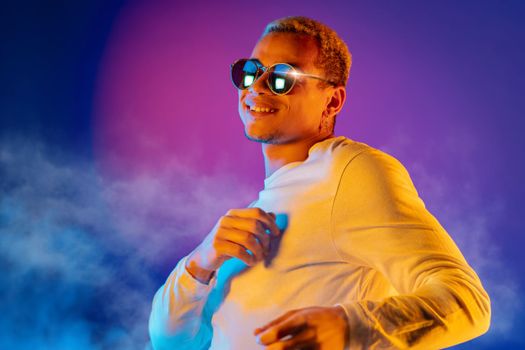 Energetic african american man dancing on colorful background under neon light. Portrait of stylish guy with dyed white curly hairstyle and round mirror eyewear. High quality photo