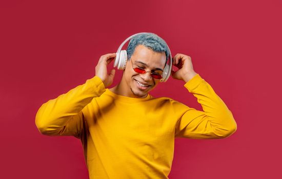 Positive american man listening music, enjoying dance with headphones on red studio background. Radio, wireless modern sound technology, online player. High quality photo