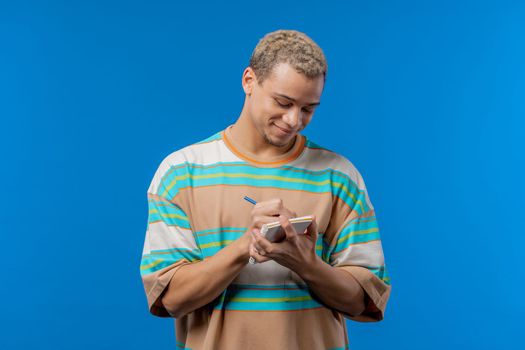 Smiling man writing notes in planner with pen. Student guy writes future plans and to-do list in notebook for week, month, year. Keeping personal diary on blue background. High quality photo