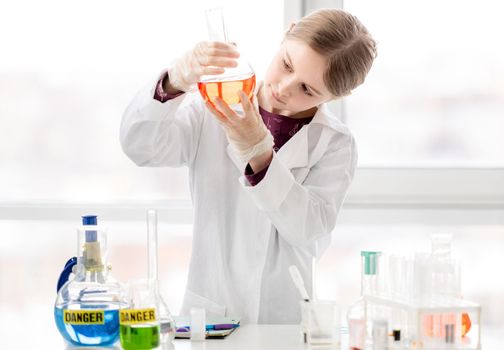 Smart girl during scientific chemistry experiment wearing protection glasses, holding bottle with orange liquid. Schoolgirl with chemical equipment on school lesson portrait