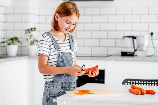 Preteen girl pilling carrot at kitchen and smiling. Child with orange vegetables food cooking at home