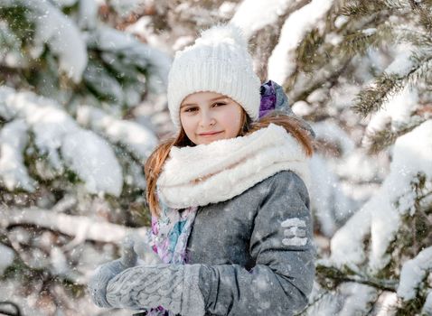 Pretty child girl in snow forest in winter time looking at camera. Preteen female kid wearing hat and gloves in cold weather with snowflakes