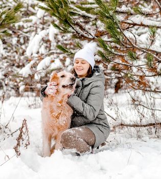 Girl petting golden retriever dog in snow forest with trees in winter time. Young woman with doggy pet outdoors in cold weather with snowflakes