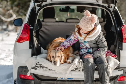 Girl child petting golden retriever dog in car trunk in winter time. Pretty kid with labrador pet doggy together in vehicle in cold weather