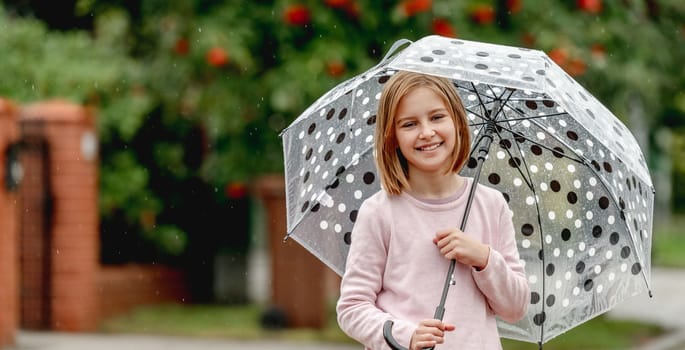 Preteen girl with umbrella looking at camera and smiling outdoors. Pretty child kid portrait in rainy day at street