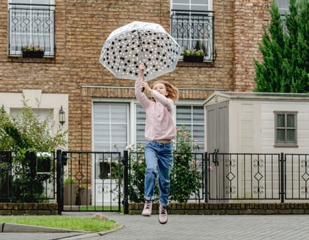 Preteen girl with umbrella running and jumping outdoors. Pretty child kid portrait in rainy day at street