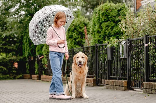 Preteen girl with golden retriever dog under umbrella at street. Pretty kid child with doggy pet in rainy day together