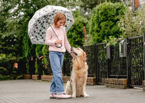 Preteen girl with golden retriever dog under umbrella at street. Pretty kid child with doggy pet in rainy day together