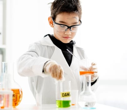 School boy wearing protection glasses with dirty face doing chemistry experiment with chemical liquids in elementary science class. Clever pupil analyzing results of test in lab