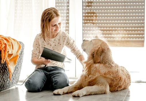 Preteen girl dries golden retriever dog wet hair with dryer after shower on floor. Pretty child kid cares about pet labrador friend indoors after bath