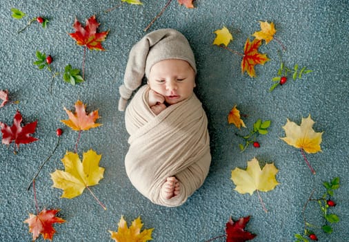 Newborn baby boy swaddled in fabric sleeping with knitted pumpkin toy and decoration. Adorable infant child kid studio halloween portrait