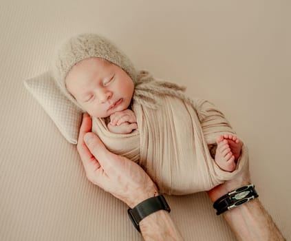 Newborn baby boy wearing knitted beige costume lying on his tummy and his father hands holding him. Adorable infant child resting and parent cares about him