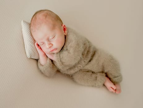 Newborn baby boy sleeping and holding hands under cheeks. Adorable infant child kid napping wearing knitted beige hat and costume