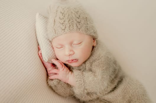 Newborn baby boy sleeping and holding hands under cheeks. Adorable infant child kid napping wearing knitted beige hat and costume