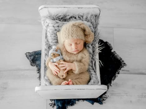 Newborn baby boy sleeping on fur in tiny bed and holding hands under cheeks. Adorable infant child kid napping on his tummy during studio photoshoot wearing knitted hat and costume