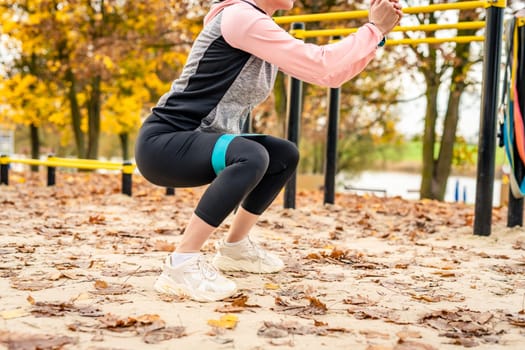 Fitness girl doing squats with eastic rubber band outdoots in autumn time. Young woman exercising in park, legs closeup