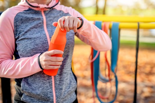Fitness girl opening bottle with water after exercising in autumn park outdoors. Young woman and hydration during training at fall season