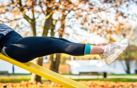 Girl holding legs up during abs ezxercises at autumn time outdoors. Young woman doing tummy training workout