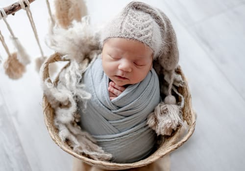 Newborn baby boy swaddled in fabric and wearing hat sleeping in basket studio portrait. Infant child kid napping