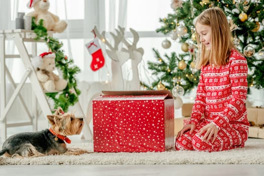 Child girl unpacks Christmas red gift box at home with cute dog. Kid celebrating New Year with presents and looking at doggy pet