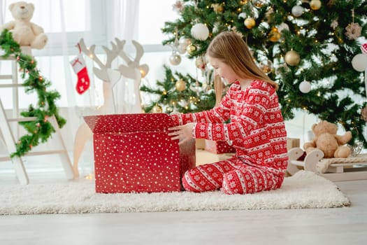 Pretty child girl open Christmas red gift box at home with traditional decorated tree. Kid celebrating New Year with presents