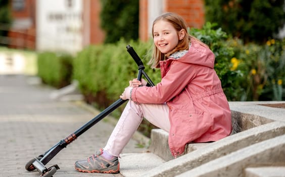 Cute litte girl sitting with a kick scooter on a street near home