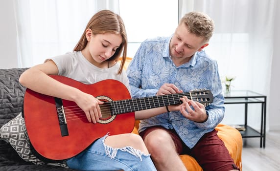 Father guy teaching girl teenager daughter guitar playing at home. Family musical lessons with strings instrument