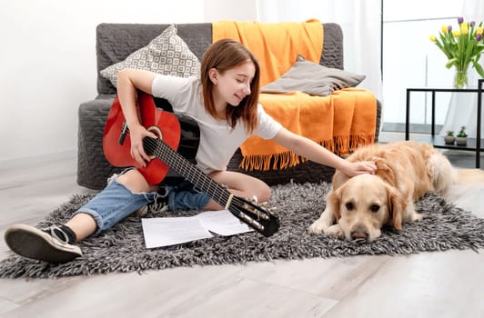 Girl teenager practicing guitar playing with golden retriever dog at home sitting on floor. Pretty guitarist with musician instrument and purebred pet doggy looking at camera