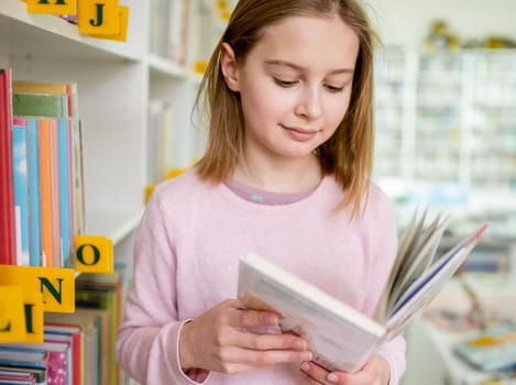 Pretty girl child reading book in library. Cute female preteen kid studying literature in biblioteque