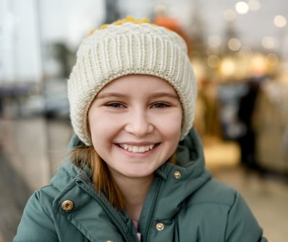 Preteen girl smiling at street portrait in city with blurred background. Cute female child kid wearing hat outdoors at autumn time