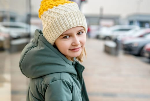 Preteen girl street portrait in city with blurred background. Cute female child kid wearing hat outdoors at autumn time