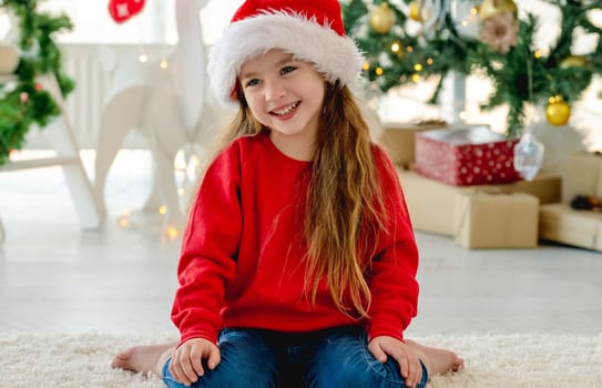 Happy child girl in Christmas time sitting on floor and smiling in room with decorated tree on background. Pretty kid wearing Santa hat at home in New Year