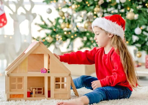 Little girl child wearing Santa hat in Christmas time sitting on floor and playing with wooden toy house at decorated New Year home with festive tree. Cute kid in Xmas time