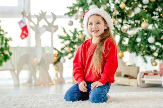 Little girl child wearing Santa hat in Christmas time sitting and smiling at decorated New Year home with festive tree. Cute kid in Xmas time