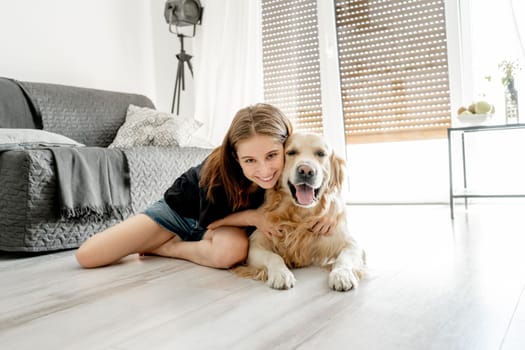 Pretty girl with golden retriever dog sitting on floor at home and hugging pet friend. Beautiful female teenager person with doggy in light room